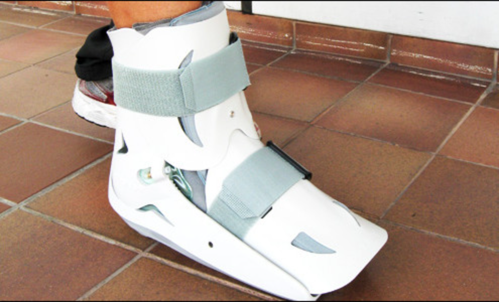 Person wearing a white ankle brace on their foot, standing on a tiled floor with a wall in the background