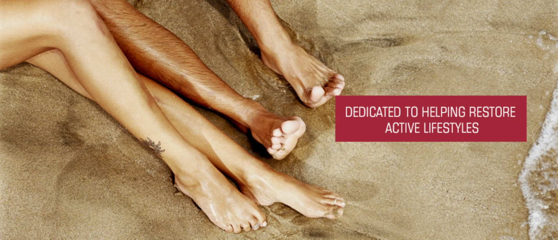 Man and woman laying on the beach with their feet in the sand, surrounded by water on the right side