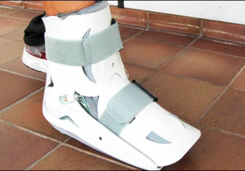 Person wearing a white ankle brace on their foot, standing on a tiled floor with a wall in the background